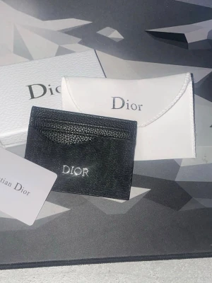 Svart Dior korthållare i läder - Snygg svart korthållare från Dior i stilrent läder med silverfärgad Dior-logga framtill. Kommer med vit fodralpåse och originalförpackning. Perfekt för att hålla koll på dina kort med stil.