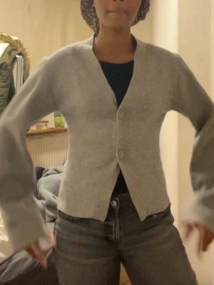 Ljusgrå cropped kofta med knappar - Snygg ljusgrå cropped kofta med två stora knappar framtill och v-ringad hals. Koftan har vida ärmar med knappdetalj vid ärmslut och är gjord i ett mjukt, tunt material. Perfekt att slänga över en topp för en chill look.