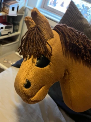 Brun käpphäst med garnman - Säljer en brun käpphäst med mjuk plysch och mörkbrun man av garn. Hästen har svarta broderade ögon och näsborrar samt två upprättstående öron. Perfekt för lek och fantasi, enkel att hålla i och använda.