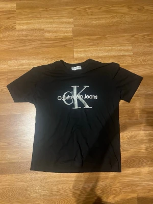 Svart Calvin Klein Jeans t-shirt - Snygg svart t-shirt från Calvin Klein Jeans med stort CK-logotyp tryckt i vitt på bröstet. Klassisk passform och rund halsringning, perfekt till jeans eller shorts. Tillverkad i mjuk bomull för skön känsla hela dagen.