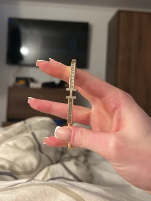 Guldigt armband med stenar - Snyggt och stilrent armband i guldton med raka linjer och två T-formade detaljer framtill. Armbandet är dekorerat med gnistrande stenar längs hela ovansidan för en lyxig känsla. Perfekt accessoar för att lyfta din outfit.