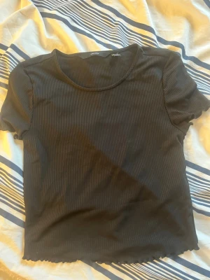 Svart ribbad croppad t-shirt - En svart ribbad t-shirt med korta ärmar och croppad passform. T-shirten har en rund halsringning och vågig kant nertill. Är använd varsamt. Material 93% polyester & 7% elastan.