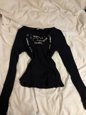 Svart ribbad topp från FB Sister - Svart långärmad topp från FB Sister med ribbad struktur och snörning framtill där det står 'OVER IT' i vitt. Figurnära passform och trendig design som sticker ut. Perfekt för dig som gillar edgy detaljer och vill ha något unikt i garderoben.