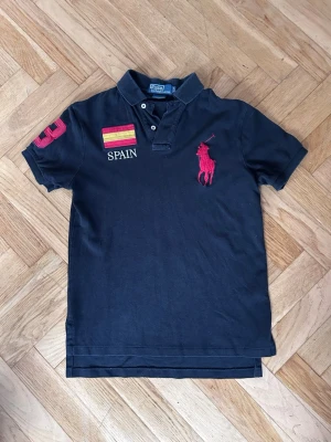 Svart pikétröja Polo Ralph Lauren - Klassisk svart pikétröja från Polo Ralph Lauren med röd logga på bröstet, broderad spansk flagga och texten SPAIN. Nummer 3 i rött på ärmen. Skön passform och krage med knappar. Perfekt för dig som gillar sportig och stilren look.