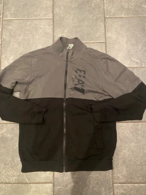 Grå och svart zip-hoodie EA7 Armani - Snygg tvåfärgad hoodie från Emporio Armani EA7 med dragkedja framtill. Överdel i grått och underdel i svart, med EA7-logga tryckt på bröstet. Tillverkad i mjukt bomullsmaterial och har ribbade muddar vid ärmslut och nederkant. Perfekt för en avslappnad streetwear-look.