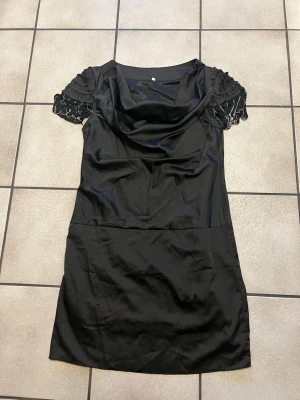 Svart kort klänning med spetsärmar storlek L - Snygg svart kort klänning med draperad urringning och unika spetsdetaljer på de korta ärmarna. Klänningen har en elegant och stilren look, perfekt för dig som Scotta sticka ut. Materialet känns mjukt och har en lätt glans.