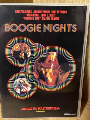 Boogie Nights DVD-film - Boogie Nights DVD med färgstark omslagsdesign och retrostil. Filmen utspelar sig i Los Angeles 1977 och har flera kända skådespelare. Omslaget har svart bakgrund med regnbågsfärgade cirklar och bilder från filmen. Perfekt för filmälskare som gillar klassiker.