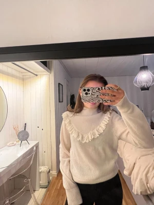 Vit stickad tröja med volang - Säljer en vit stickad tröja med hög hals och dekorativ volang runt halsen. Tröjan har långa ärmar och är gjord i ett mjukt material som känns skönt mot huden. Perfekt för dig som gillar en söt och trendig look.