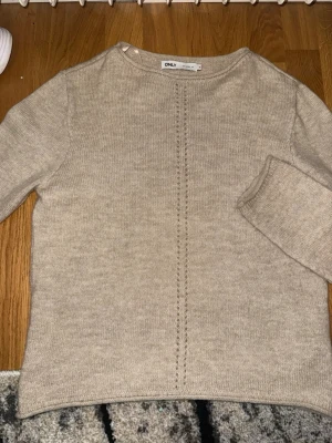 Beige stickad tröja från ONLY - Säljer en beige stickad tröja från ONLY med rund halsringning och dekorativ hålstickad detalj mitt fram. Tröjan har långa ärmar och är gjord i mjukt material som känns skönt mot huden. Enkel och stilren design som passar till det mesta.