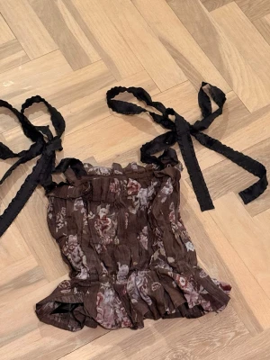Brun blommig linnetopp med knytband - Supersöt brun linnetopp med blommigt mönster och breda svarta knytband på axlarna. Toppen har volangkant nedtill och en lätt rynkad struktur som ger en romantisk vibe. Perfekt att styla med jeans eller kjol för en trendig look.