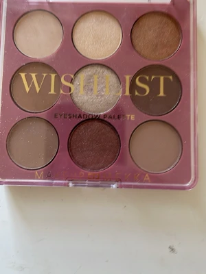 Wishlist ögonskuggspalett Makeup Mekka - Wishlist Eyeshadow Palette från Makeup Mekka med nio nyanser i jordnära toner som beige, brunt, champagne och guld. Paletten har både matta och skimriga skuggor i rund form, perfekt för att skapa olika ögonlooks. Förpackningen är rosa-aktig med genomskinligt lock och guldfärgad text.