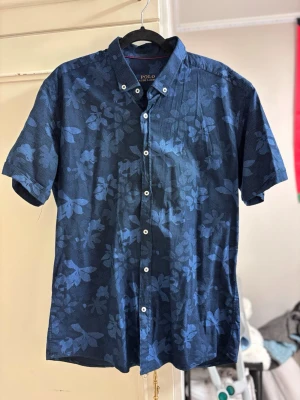Polo Ralph Lauren skjorta  - Snygg blå skjorta från Polo Ralph Lauren med korta ärmar och blommigt mönster. Skjortan har klassisk krage, vita knappar och broderad logga på bröstet. Storlek L men passar mer M/S (längd: 68cm, bredd: 50 cm. Perfekt nu för sommaren 