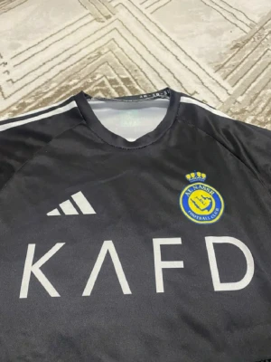 Svart Al Nassr fotbollströja Adidas - Säljer en svart Al Nassr fotbollströja från Adidas med klubbens emblem på bröstet och vita detaljer. Tröjan har tryck med 'KAFD' på framsidan och 'RONALDO 7' på ryggen. Materialet är lätt och andas, perfekt för träning eller match.