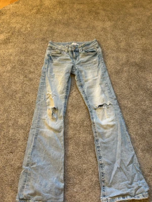 Ljusblå slitna jeans från Gina Tricot - Ljusblå jeans från Gina Tricot med slitningar och hål på båda knäna. Modellen har utsvängda ben och klassisk femficksdesign. Perfekta för en avslappnad och trendig look. Tillverkade i denim med snygg tvätt.