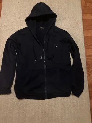 Navy blue hoodie från Polo Ralph Lauren - Säljer en svart hoodie med dragkedja från Polo Ralph Lauren. Klassisk design med huva och dragsnören, samt den ikoniska lilla vita logotypen broderad på bröstet. Tillverkad i mjukt bomullsmaterial och har två fickor framtill.
