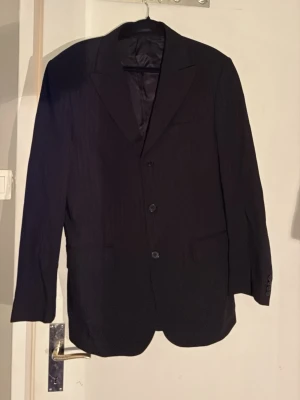 Svart klassisk kavaj - En svart kavaj med klassisk passform och tre knappar framtill. Kavajen har breda slag. Perfekt för en stilren look och passar till skjorta eller t-shirt. Materialet ser ut att vara en blandning av polyester och viskos.