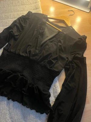 Svart omlottopp med spetsdetaljer från  - Trendig svart omlottopp med v-ringning och spetsdetaljer längs urringningen. Toppen har långa ärmar, smockad midja och volangkant nedtill för en snygg siluett. Perfekt att styla med jeans eller kjol.