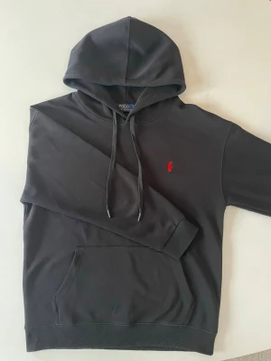 Svart hoodie från Polo Ralph Lauren - Snygg svart hoodie från Polo Ralph Lauren med klassisk röd broderad logga på bröstet. Hoodien har huva med dragsko, magficka och ribbade muddar. Tillverkad i mjukt bomullsmaterial som är skönt att bära. Perfekt för en avslappnad och stilren look.
