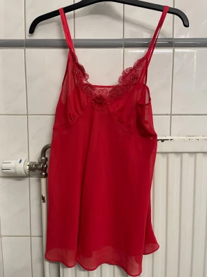 Rött nattlinne med spetsdetaljer - Snyggt rött nattlinne med tunna axelband och transparent tyg. Ovanför bysten finns fina spetsdetaljer och broderade blommor som ger en romantisk känsla. Nattlinnet har en lös och luftig passform, perfekt för mysiga kvällar.