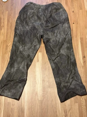 Vida svarta byxor i mockaimitation - Säljer ett par vida byxor i svart mockaimitation med raka ben och två bakfickor. Byxorna har en cool vintage-look tack vare materialet och den matta finishen. Perfekta för dig som vill ha en unik stil och gillar retrovibbar.