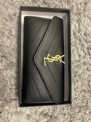 Svart plånbok YSL - Snygg svart plånbok från Saint Laurent i äkta läder med guldfärgat YSL-emblem på framsidan. Plånboken har ett elegant kuvertformat lock och flera kortfack på insidan. Perfekt för dig som vill ha en lyxig accessoar med ikonisk design.