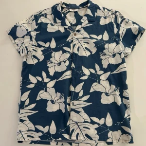 Blå blommig skjorta från KappAhl - Kortärmad skjorta från KappAhl i mörkblått med stort vitt blommigt mönster. Skjortan har klassisk krage, knappar framtill och är tillverkad i ett lätt och svalt material, perfekt för varma dagar.