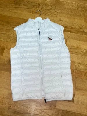 Vit dunväst från JOTT - Snygg vit dunväst från JOTT med quiltad design och dragkedja framtill. Västen har två sidofickor med dragkedja och en liten logotyp på bröstet. Även två fickor intill västen. Perfekt för lager-på-lager och stilren look.