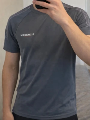 Mörkgrå McKenzie t-shirt - Snygg mörkgrå t-shirt från McKenzie med logga på bröstet och diskreta detaljer längs axlarna. T-shirten har en klassisk passform och är tillverkad i ett mjukt syntetmaterial som passar perfekt till träning eller chill outfits. Mycket bra skick och storlek: S