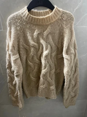 Beige kabelstickad tröja från NA-KD - Säljer en beige kabelstickad tröja från NA-KD med grova flätmönster framtill och på ärmarna. Tröjan har rund halsringning, ribbade muddar och är gjord i ett mjukt, fluffigt material.