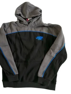 Svart och grå hoodie med blå detaljer - Snygg hoodie i svart och grått med blå piping längs ärmar och bröst. Hoodien har en huva och en liten blå logga på bröstet. Tillverkad i mjukt material, perfekt för en avslappnad streetwear-look.