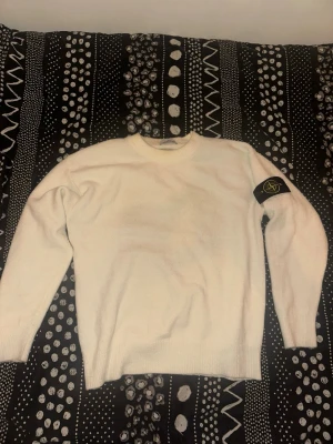 Vit stickad tröja från Stone Island - Vit stickad tröja från Stone Island med klassisk rund halsringning och den ikoniska svarta logotypen med gult och grönt på vänster ärm. Tröjan har ribbade muddar vid ärmslut och nederkant. Perfekt för dig som gillar stilrena plagg med streetwear-vibe. 