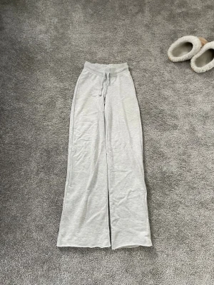 Grå mjukisbyxor från Gina Tricot - Säljer ett par ljusgrå mjukisbyxor från Gina Tricot i storlek XS. Byxorna har raka, vida ben och resår i midjan med dragsko för skön passform. Perfekta för chill dagar hemma eller när du vill vara extra bekväm.