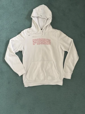 Vit hoodie från Puma med rosa tryck - Snygg vit hoodie från Puma med ett rosa mönstrat logotyptryck på bröstet. Hoodien har huva med dragsko och en stor magficka framtill. Tillverkad i mjukt material och har ribbade muddar vid ärmar och nederkant. Perfekt för en chill och sportig look.