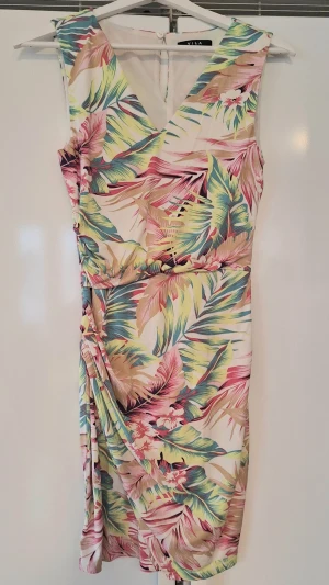 Blommig kortklänning från Vila S - Snygg kortklänning från Vila med tropiskt blommigt mönster i rosa, grönt, beige och blått. Klänningen har v-ringning, omlott-detalj i kjolen och är ärmlös. Perfekt för sommarens alla tillfällen! Stretchig.