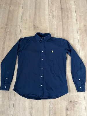 Marinblå skjorta från Polo Ralph Lauren - Klassisk marinblå skjorta från Polo Ralph Lauren med broderad gul logga på bröstet. Skjortan har knappar framtill och på ärmsluten samt en stilren krage. Perfekt för en stilren look.
