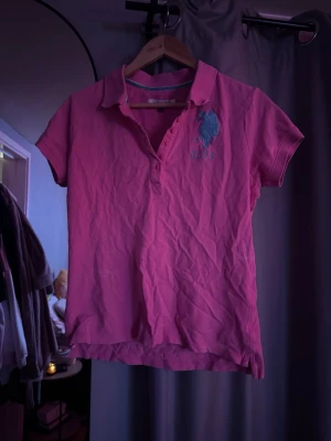 Rosa pikétröja från US Polo Assn - Säljer en rosa pikétröja från US Polo Assn med klassisk krage och knappar framtill. Tröjan har korta ärmar och ett stort blått logotryck på bröstet. Storlek L men skulle säga passar som en M