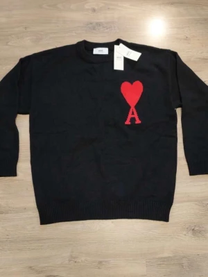 Svart stickad tröja från Ami Paris - Svart stickad tröja från Ami Paris med rund halsringning och ribbade muddar. På bröstet finns ett rött hjärta med bokstaven A, vilket är varumärkets ikoniska logga. Perfekt för dig som gillar stilrena och trendiga plagg.