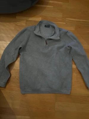 Grå halvzip tröja från Polo Ralph Lauren - Snygg grå halvzip tröja från Polo Ralph Lauren med klassisk broderad logga på bröstet. Tröjan har hög krage, dragkedja och är tillverkad i mjukt bomullsmaterial. Perfekt för lager på lager och chill stil.