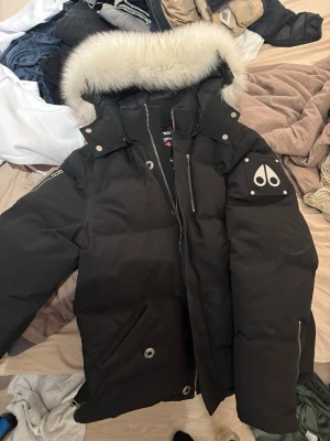 Svart Moose Knuckles jacka  - Säljer en svart dunjacka från Moose Knuckles äkta från moose knuckels med stor vit pälskant på huvan och ikonisk logga på ärmen. Jackan har flera fickor, dragkedja och tryckknappar framtill samt detaljer i metall. Perfekt för kalla vinterdagar.