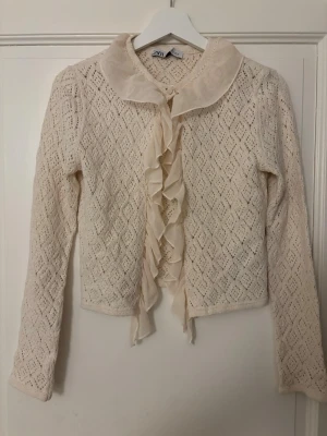 Krämvit spetskofta från Zara - Söt krämvit kofta från Zara med spetsmönster och volangkant framtill. Koftan har lång ärm, dekorativ krage och är tillverkad i ett tunt, virkat material. Perfekt för en romantisk och vintage-inspirerad look.