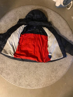 Svart pufferjacka från Gant - Säljer en svart pufferjacka från Gant med röd och grå insida. Jackan har huva och tryckt text på insidan med 'GANT 1949 NEW HAVEN, CONNECTICUT U.S.A.'. Perfekt för kalla dagar och riktigt snygg streetstil.