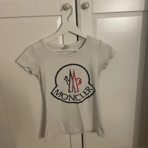 Vit Moncler t-shirt  - Vit t-shirt från Moncler med stor ikonisk logga i svart och rött framtill. Klassisk rund halsringning och korta ärmar. Perfekt för dig som gillar stilrena och trendiga plagg med tydlig branding. Har samma i svart skriv om du vill ha bild på den okså