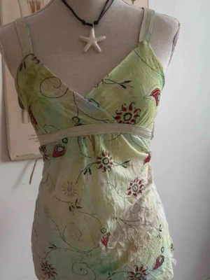 Broderad omlottopp i grönt och rött - Snygg omlottopp med broderade blommor i grönt, rött och beige. Toppen har breda axelband, omlottskärning och ett band under bysten. Materialet är tunt och lätt, perfekt för varmare dagar.