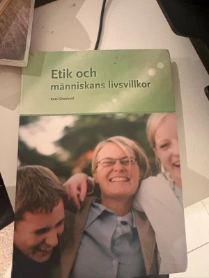 Etik och människans livsvillkor - Kurslitteratur med grönt omslag och titeln 'Etik och människans livsvillkor' av Katri Cronlund. Boken har ett mjukt omslag med motiv av människor i bakgrunden och används ofta inom vård- och omsorgsutbildningar.