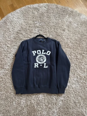  Polo Ralph Lauren sweatshirt  - Snygg mörkblå sweatshirt från Polo Ralph Lauren med vit text 'POLO R-L' och ett tigertryck på bröstet. Tröjan har rund halsringning, långa ärmar och ribbade muddar vid ärmslut och nederkant. Perfekt för en avslappnad och sportig stil.
