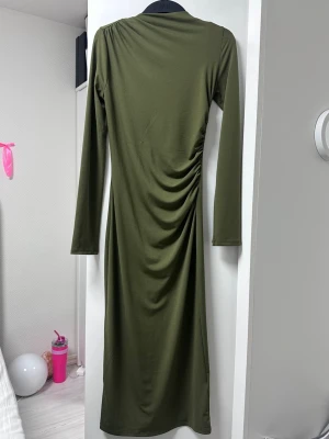 Olivgrön långärmad klänning - Snygg olivgrön långklänning med lång ärm och draperad detalj på sidan. Klänningen har en enkel, rak passform och är tillverkad i ett mjukt, stretchigt material som ger en bekväm känsla.