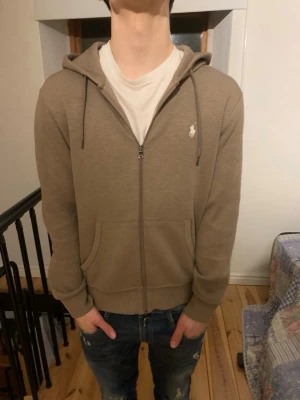 Beige hoodie från Ralph Lauren - Snygg beige hoodie med dragkedja från Ralph Lauren. Klassisk design med huva, dragsnören och två fickor framtill. Liten vit logga på bröstet. Perfekt för en avslappnad och stilren look. Den är i väldigt bra skick men är lite sliten runt dragkedjan.