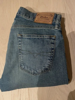 Blå raka jeans från Polo Ralph Lauren - Snygga blå jeans från Polo Ralph Lauren med klassisk femficksdesign och raka ben. Jeansen har en läderpatch med logga bak i midjan och subtila slitningar för en avslappnad look. Perfekta till vardagsstilen och tillverkade i bomull.