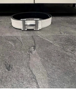 Vitt Hermès bälte med silver H-spänne - Säljer ett vitt bälte från Hermès med det ikoniska H-spännet i silverfärgad metall. Bältet är gjort i skinn och har en stilren, klassisk design som passar till många olika outfits. Perfekt accessoar för dig som gillar lyxiga detaljer.
