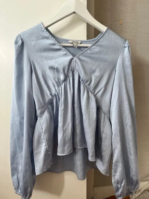 Ljusblå volangblus från AWARE - Supersöt ljusblå blus från AWARE med volangdetaljer framtill och v-ringning. Blusen har lång ärm med lätt puff och är tillverkad i ett glansigt, tunt tyg som ger en elegant känsla. Perfekt för dig som gillar en feminin och trendig stil.